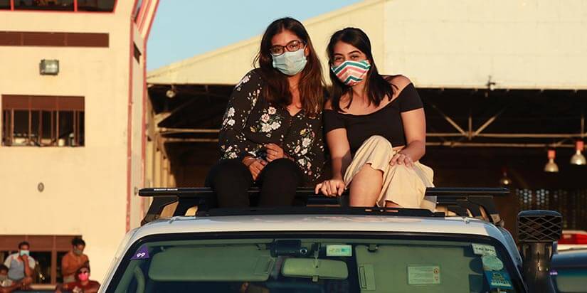 Dua orang wanita duduk di atas mobil sambil mengenakan masker kain